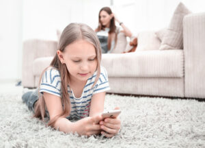 Child using a smartphone on a living room rug while a parent relaxes on the sofa nearby